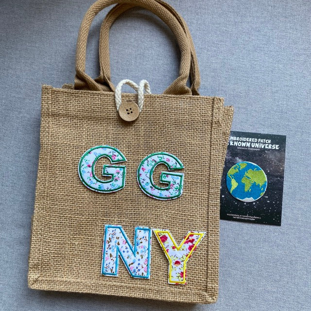 floral letter embroidered patches spelling GGNY on a burlap tote bag with product packaging card