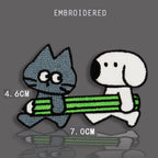 grey tabby cat and white Snoopy-style dog carrying a green striped pencil together one at each end embroidered stick-on patch 70x46mm