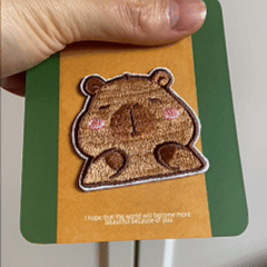 animated hand holding packaged derpy capybara embroidered patches showing two styles and detailed embroidery texture