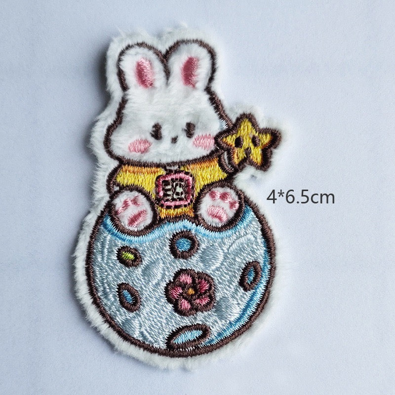 bunny sitting on blue spotted planet embroidered stick-on patch — white bunny upright in gold spacesuit with yellow star beside floral planet surface 40x65mm