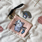 Fishing cat embroidered stick-on patch on a pink pouch beside a cat carrying a large fish patch