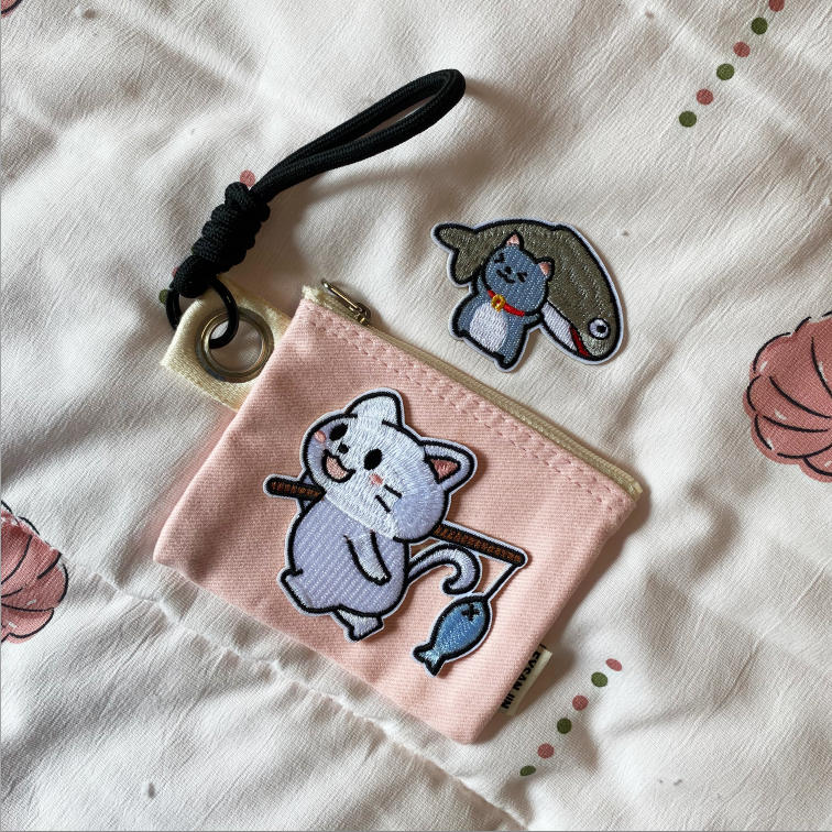 Fishing cat embroidered stick-on patch on a pink pouch beside a cat carrying a large fish patch