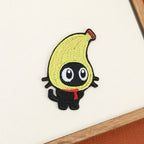 Playful black cat embroidered stick-on patch with a yellow banana hood and bright red scarf
