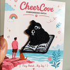 animated hand holding packaged funny black kitty void reading book embroidered patch showing detailed embroidery texture